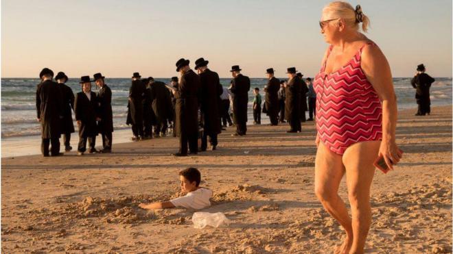 Uma mulher de maiô observa um grupo de judeus ortodoxos em uma praia perto de Tel Aviv