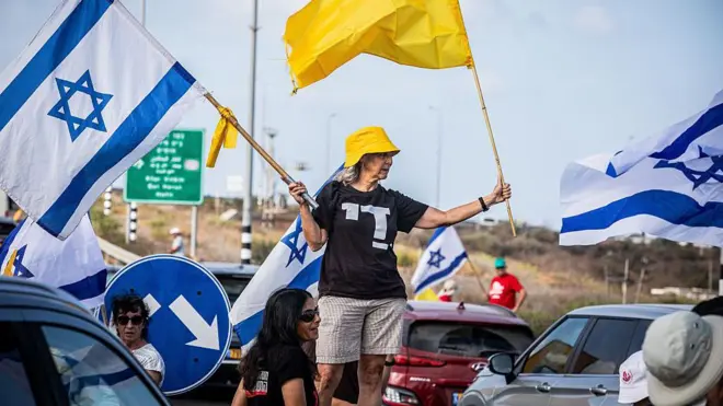 Une femme tenant un drapeau israélien et un drapeau jaune représentant les otages les agite tout en se tenant au milieu d'une route très fréquentée lors d'une manifestation diurne.