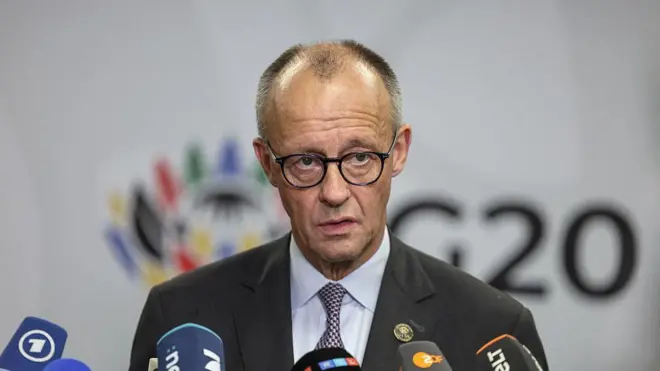 Merz takım elbiseli ve mikrofonların olduğu kürsüde ayakta konuşuyor. 22 Kasım 2025. 