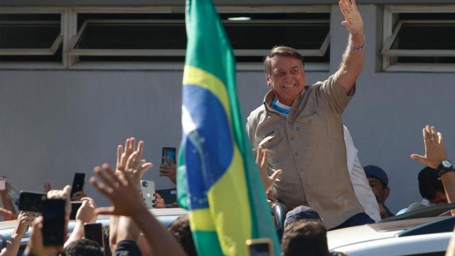 Bolsonaro sorrindo acena para apoiadores dianteroleta de clubes de futebolbandeira do Brasil