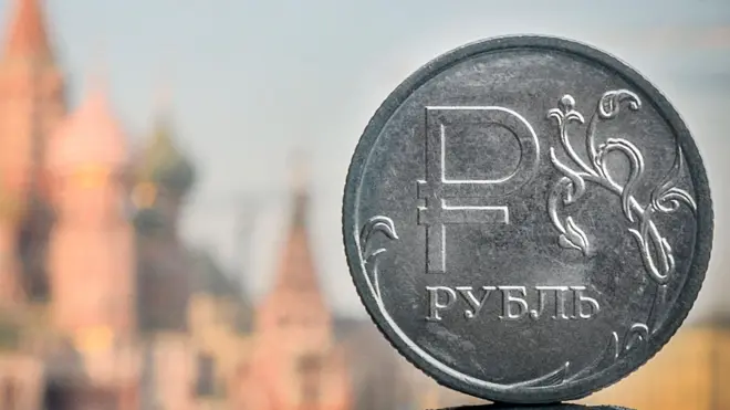 На заднем плане размытые очертания православного собора Василия Блаженного в Москве. На переднем плане монета из серого металла с надписью "рубль"
