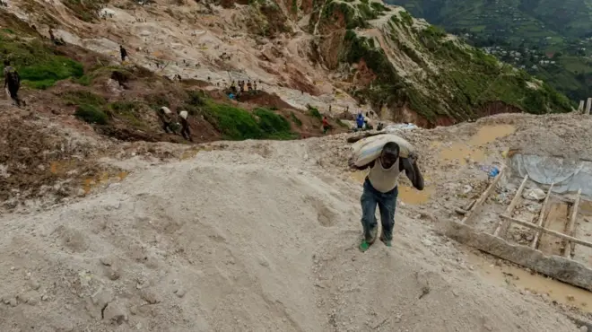 Un mineur transporte un sac de minerai à la mine de Rubaya. Le terrain est vallonné. Derrière lui, on aperçoit d'autres mineurs et, au loin, des champs verdoyants. 