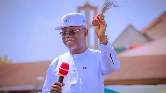 Aworan Gomina Lucky Aiyedatiwa pẹlu ẹrọ gbohungbohun lọwọ ọtun rẹ, igbalẹ idanimọ ẹgbẹ oṣelu APC lọwọ osi lasiko ipolongo ibo kan.