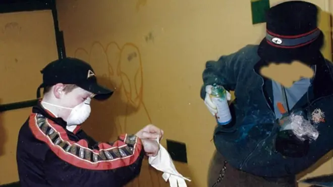 Банксy леанс то оне сиде холдинг а спраy цан, wхиле тwо цхилдрен пут он маскс анд гловес.