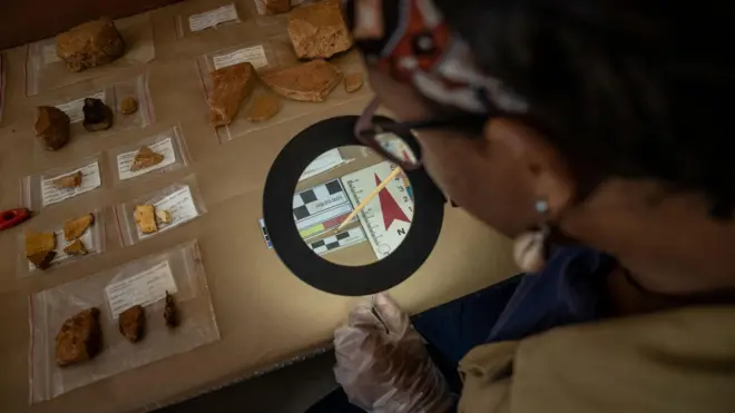 Foto de uma mulher olhando por uma espécie de lupa material arqueológico encontrado. 