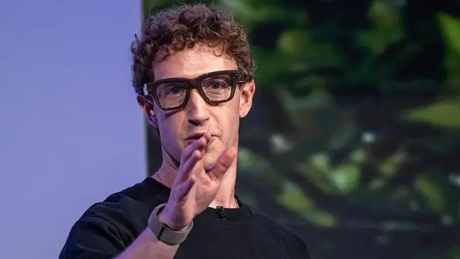 Mark Zuckerberg wearing thick black rimmed smart glasses and gesturing with his hand.