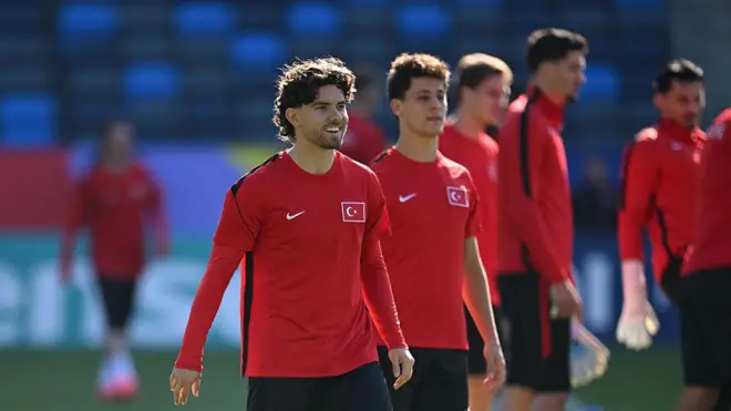 Ferdi Kadıoğlu sahada gülümsüyor. Arkasında bir dizi Türk milli futbolcu var