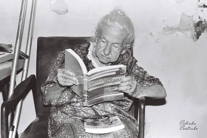 Cora Coralina sentada numa cadeira lendo