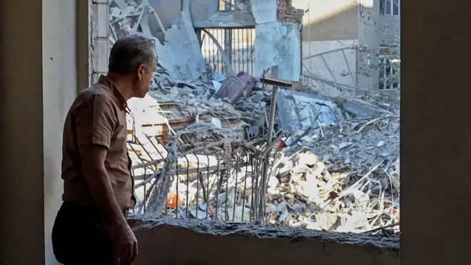 مردی از درون اتاقی که پنجره‌اش ویران شده به ویرانه‌های بیرون نگاه می‌کند