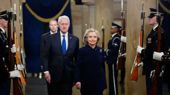 Билл и Хиллари Клинтон идут по коридору, вдоль которого стоят военные с винтовками. Оба одеты в темные костюмы.