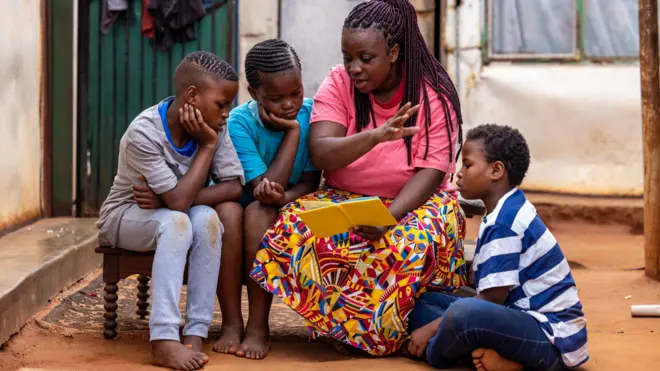 Foto of mother wey dey read book wit her three children.