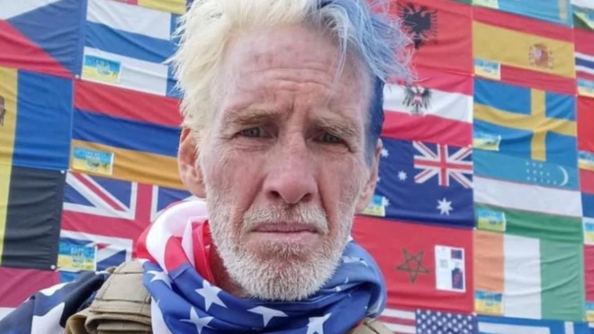 Сивий чоловік у військовій жилетці та шарфі з американським прапором. Позаду нього як фон великий візерунок з прапорів.