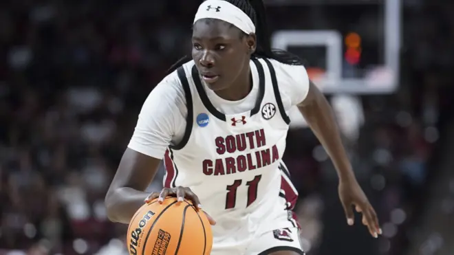 Basketballer Madina Okot dribbles an orange ball forward on an indoor court. The player wears a white South Carolina jersey with the number 11 on the front and a white headband, leaning slightly forward in motion. A hoop and blurred spectators are visible in the background under bright arena lighting