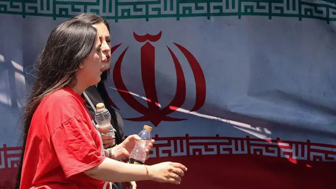 Tahran'da arkada İran bayrağı görünen bir sokakta iki kadın yürüyor. Kadınların başı açık ve biri kırmızı tişört giyiyor.