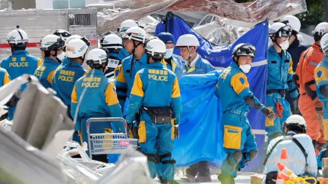 がれきの散乱する中、さまざまな熊本県警などさまざまな所属の隊服を着た人たちたヘルメットやマスクをつけて立っている