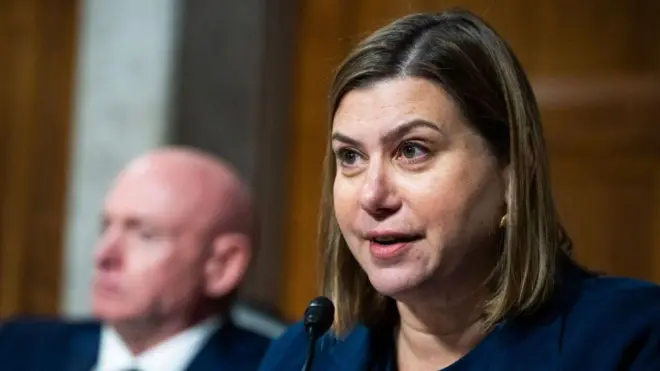 青いスーツを着た、ブロンドをボブカットにした女性が、マイクに向かって話している。背後にはスーツを着た男性の姿がぼやけて写っている