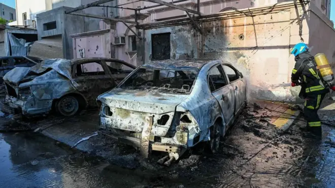 Un bombero echa agua con una manguera en dirección de unos autos quemados por la caída de esquirlas de drones
