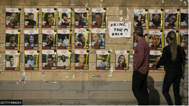 إسرائيليان يمران قرب ملصقات لصور الرهائن المحتجزين في غزة معلقة على حائط في تل أبيب مع لافتة تقول "أعيدوهم"