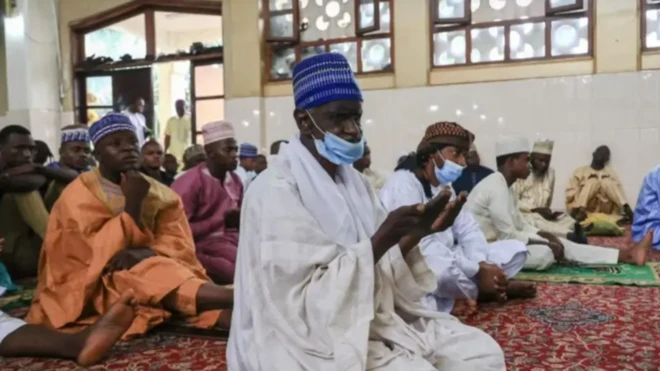 Olujọsin kan wọ agbada funfun, o de fila buluu niwaju awọn olujọsin mii tawọn naa wọ oriṣiiriṣii aṣọ buba