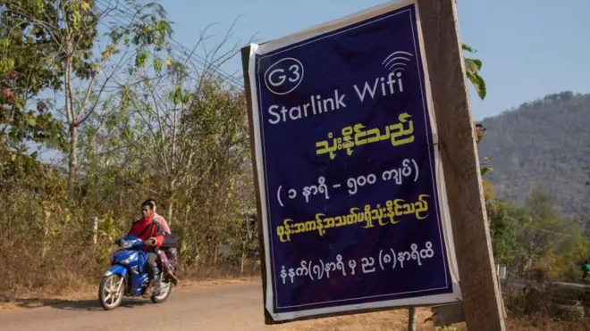 在缅甸克耶邦，一块贴在木板上的纸质告示以英文写着“Starlink Wifi”，并附有缅文说明。背景中可见两人骑着一辆蓝色电单车行驶在道路上，远处则是绿树覆盖的山丘。（16/2/2025）