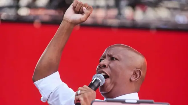 Julius Malema wear white, e raise one of im hand up as a symbol of solidarity, e use di moda hand hold microphone as e open mouth dey tok. Di background na red wit blur backgorund wey resemble im supporters.