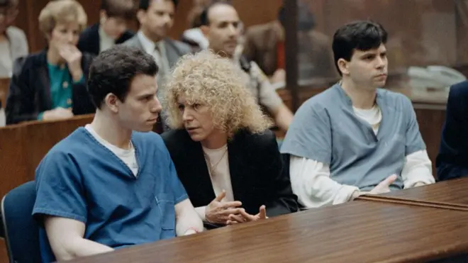 Di Menendez brothers, wearing different shades of a blue prison uniform, sit at a table wit one of dia attorneys during dia trial for 1994. Inside di courtroom, dia lawyer na woman wit curly hair and appear deep in conversation wit Erik Menendez. 