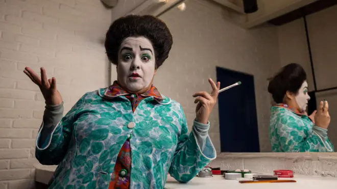 Gabriela Muñoz en un camerino, con un pincel en una mano, muestra a la cámara su maquillaje del personaje Chula: con una ceja levantada, un corazoncito pintado en los labio y un grueso pelo crespo. Un espejo la refleja de ángulo.