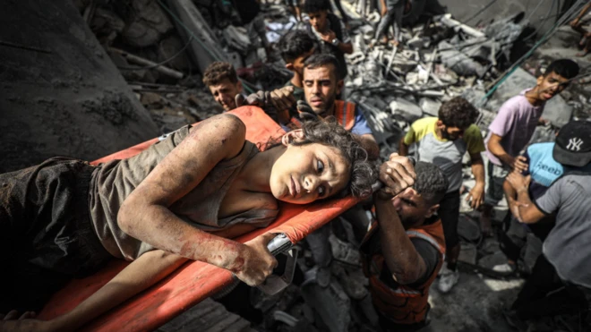 Genç bir kız, acil durum görevlileri tarafından yıkılmış bir binadan çıkarılırken sedye üzerinde yatıyor. Sedyenin kenarına tutunuyor ve üzgün görünüyor.