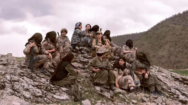 Female PJAK fighters holding their weapons sit on a rocky hilltop. Some wear head and face coverings and many carry weapons.