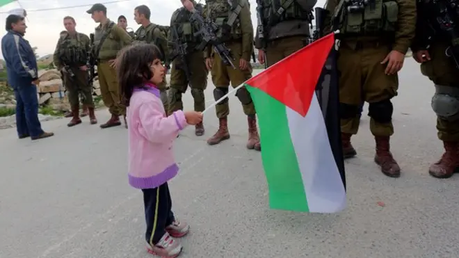 A Palestinian girls waves the Palestinian flag near Israeli soldiers during a protest over Israeli occupation and land confiscation in the West Bank village of Masarah, 19 December 2014, near the town of Bethlehem, where Jesus Christ was born
