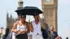 Two young women wearing white sleeveless tops and sunglasses walk down the street under a black umbrella in front of Big Ben
