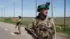 На дорозі серед відкритої місцевості стоїть військовий у тактичному спорядженні, тримаючи в руках рушницю. Позаду видно ще одного озброєного військового. Над дорогою натягнута велика протидронова сітка, закріплена на високих металевих опорах. Обабіч дороги простягаються поля та зелені насадження.