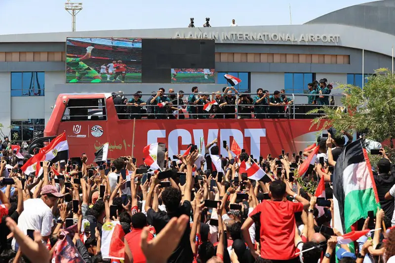 لاعبو المنتخب المصري يلوحون بأيديهم من على متن حافلة مكشوفة، وسط حشود من المشجعين الذين تجمعوا لاستقبالهم في مطار العلمين الدولي بمدينة العلمين الساحلية الشمالية في 10 يوليو/تموظ عام 2026. ويظهر مشجعون يلوحون للاعبين ويحملون أعلام مصرية، ويحاول بعضهم التقاط صور بهواتفهم المحمولة.