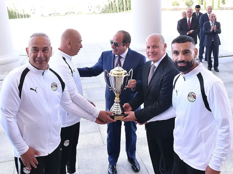 الرئيس المصري، عبد الفتاح السيسي، يحمل كأساً، بينما يربت على كتف حسام حسن، مدرب المنتحب المصري الوطني، وحوله بعض من لاعبي المنتخب المصري ومسؤولون.