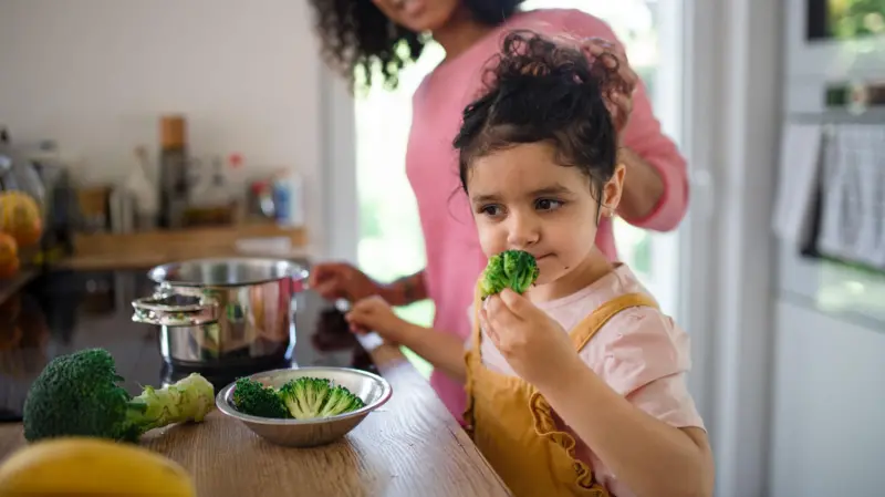 طفلة تقف في المطبخ أمام طاولة تشتم البروكلي