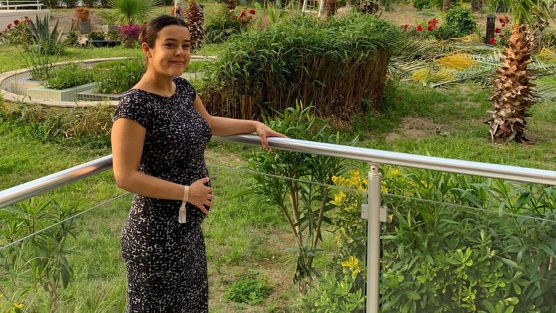 Tallulah Clark posa de pie para la foto con una mano sobre su barriga de embarazada y un jardín de fondo
            