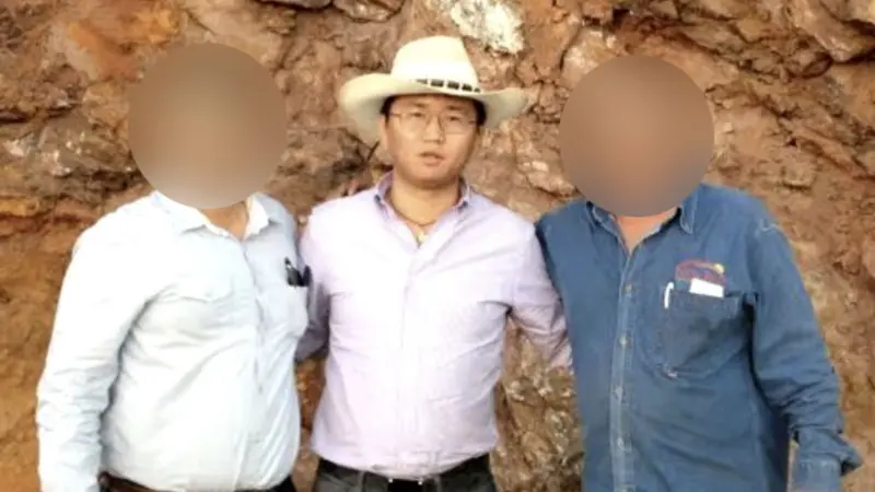 رجل يرتدي قبعة بيضاء على طراز رعاة البقر وقميصاً بنفسجياً باهتاً مدسوساً في بنطاله. يرتدي نظارة وقلادة وينظر إلى الكاميرا. يوجد رجل على يساره وآخر على يمينه - تم تشويش وجهي الرجلين لإخفاء هويتهما.