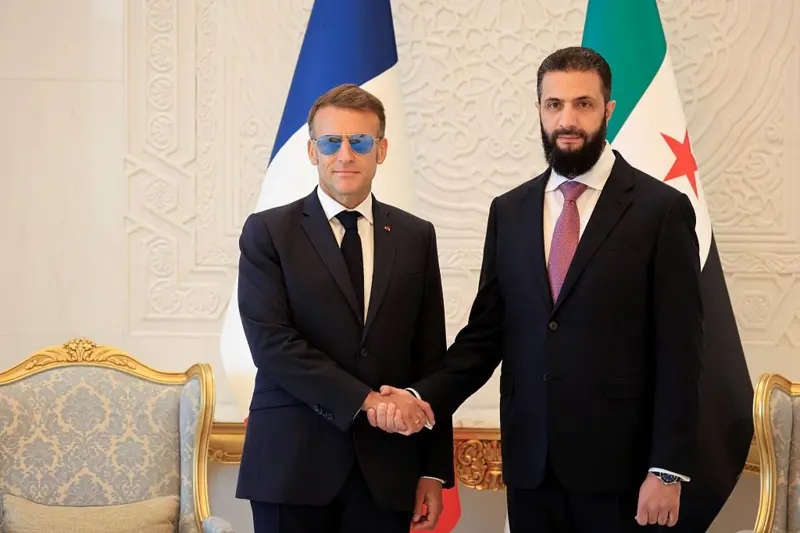 الرئيس الفرنسي إيمانويل ماكرون (يساراً)، يصافح الرئيس السوري أحمد الشرع قبيل اجتماعهما في "قصر الشعب" بدمشق في 7 يوليو/تموز 2026. ويرتدي الشرع سترة سوداء وقميصاً أبيض وربطة عنق حمراء داكنة وتغطي وجهه لحية سوداء، بينما يرتدي ماكرون سترة سوداء وقميصاً أبيض وربطة عنق سوداء، ونظارة شمس.