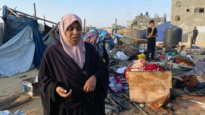 Une Palestinienne regarde l'appareil photo alors qu'elle inspecte les dégâts subis par un camp de personnes déplacées à Khan Younis, après une attaque israélienne le 2 juillet. D'autres personnes, des tentes brisées et des objets éparpillés sont visibles à l'arrière-plan, dans un ciel bleu.