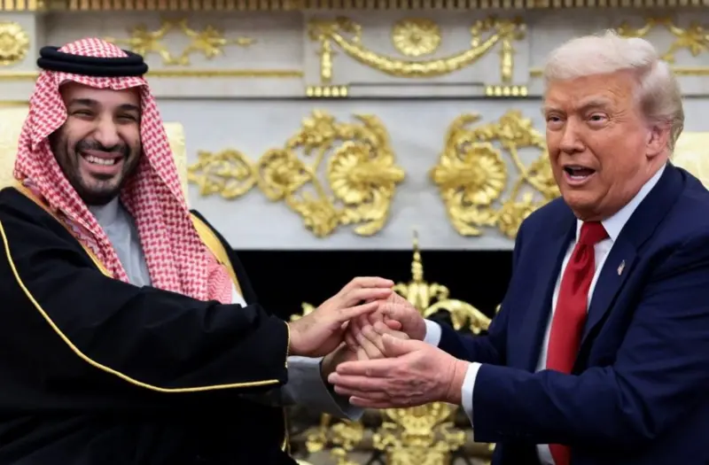 الرئيس الأمريكي دونالد ترامب وولي العهد السعودي ورئيس الوزراء محمد بن سلمان يمسكان بأيدي بعضهما خلال اجتماع في المكتب البيضاوي في البيت الأبيض في واشنطن العاصمة. يرتدي ترامب بدلة رسمية كحلية اللون وقميصاً أبيض وربطة عنق حمراء، يرتدي بن سلمان اللباس التقليدي عباءة سوداء وثوباً أبيض.