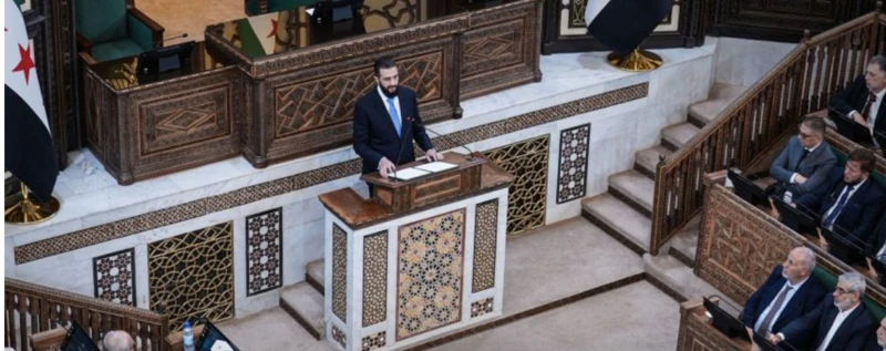 الرئيس السوري المؤقت، أحمد الشرع، يلقي كلمة خلال جلسة مجلس الشعب الأولى منذ سقوط الأسد، وذلك في العاصمة السورية دمشق، ويرتدي الشرع سترة سوداء وقميصاً أبيض وربطة عنق لونها أزرق فاتح، بينما يقف متحدثاً من على منبر ويجلس أمامه أعضاء مجلس الشعب.