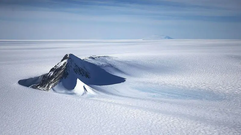 Foto de un paisaje de la Antártica en el que sobresale una colina de color netro en medio del terreno blanco