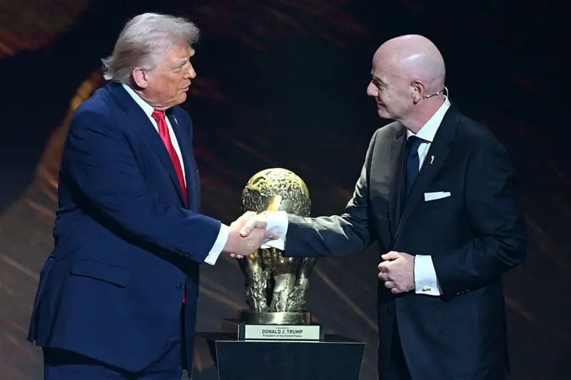 الرئيس الأمريكي دونالد ترامب يصافح رئيس الاتحاد الدولي لكرة القدم (ألفيفا) جياني إنفانتينو أثناء تسلمه "جائزة الفيفا للسلام"، وذلك خلال مراسم سحب قرعة بطولة كأس العالم لكرة القدم 2026، ويرتدي ترامب سترة زرقاء وقميصاً أبيض وربطة عنق حمراء، بينما يرتدي إنفانتينو سترة سوداء وقميصاً أبيض وربطة عنق زرقاء.