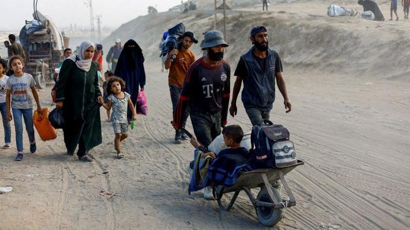 Hombres, mujeres y niños caminan con sus pertenencias sobre una zona arenosa. Un hombre empuja una pequeña carretilla donde está un niño con un bolso y pertenencias 