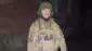 迷彩柄の軍服にヘルメットをかぶった男性を、暗闇の中でフラッシュ撮影した写真。男性はひげを伸ばしており、軍服には複数のナイフが装着されている。また、男性の背後にはれんが造りの建物が見える。