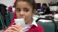 幼い女の子が、プラスチック製の肺活量測定器に息を吹き込んでいる。赤いリボンと赤いカーディガン、白いシャツを着て、学校で緑色の椅子に座っている。
