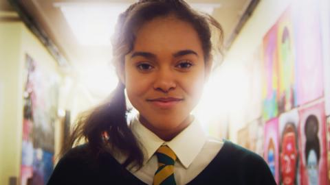 A girl in school uniform in a corridor smiling as she is surrounded by bright light, Polly.