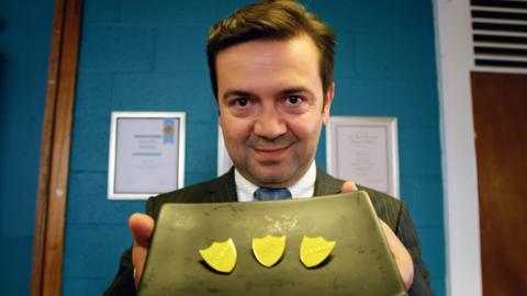 A man in a suit holding up a tray of prefect badges.