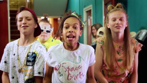 Four school kids dressed in their own bright clothes in a school corridor rapping about a competition.