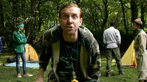 A man looks surprised and scared as people stand behind him inside a ring of tents.